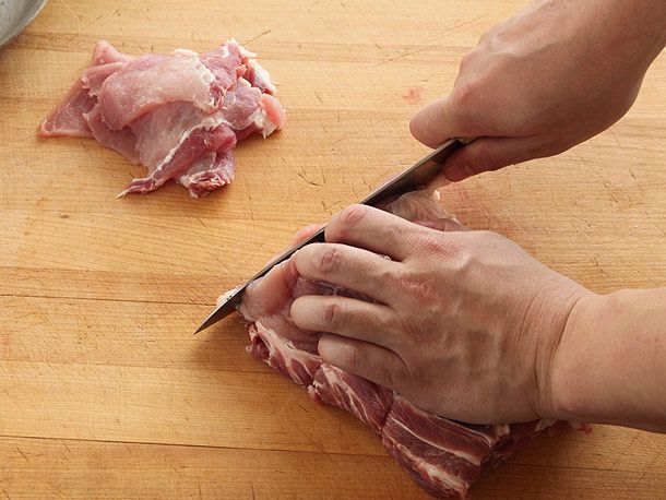 Two hands slicing raw pork sirloin against the grain on a wooden surface