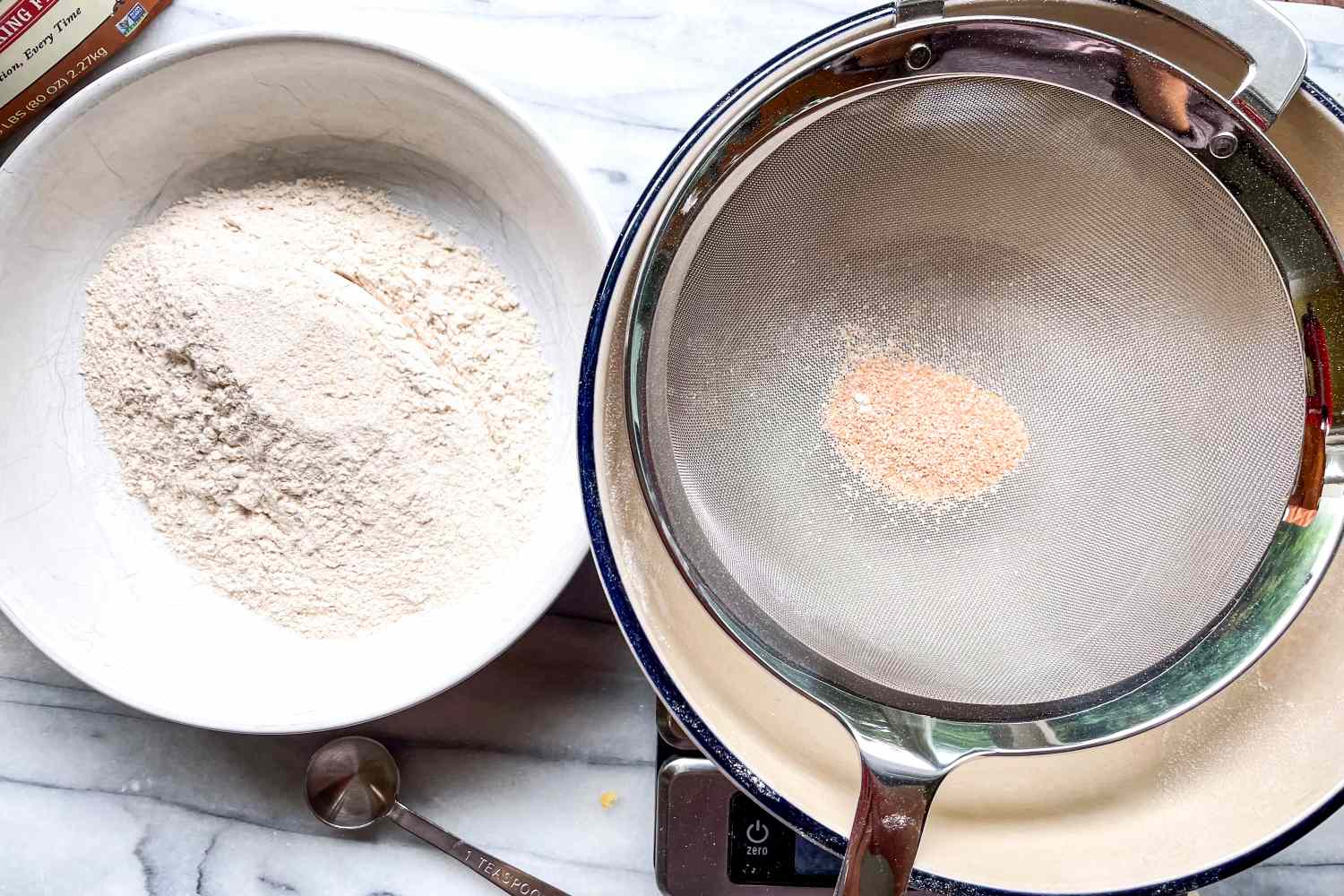 A strainer over a mixing bowl next to a bowl of sifted flour on a countertop