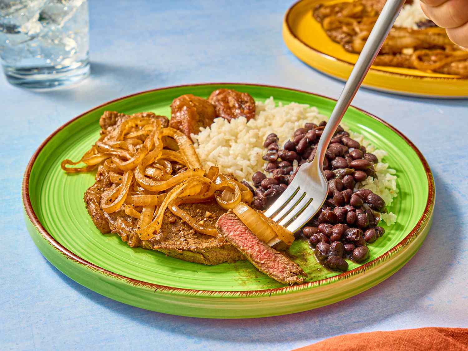 Green dish of pallomilla with rice and beans. A fork has a pink piece of steak on it with some grilled onion 