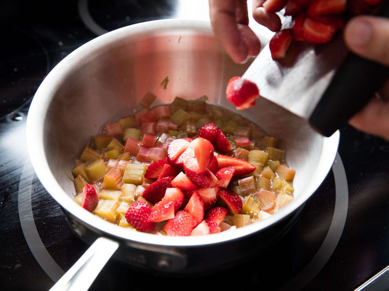 Strawberries being added to a pan containing rhubarb simmering in liquid.