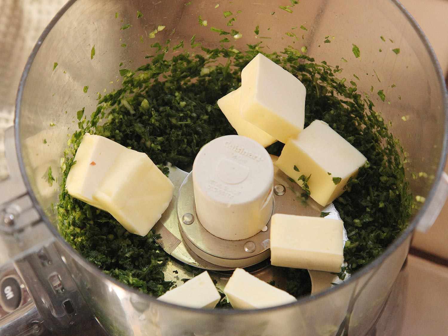 A food processor containing finely chopped green herbs with butter chunks on top.