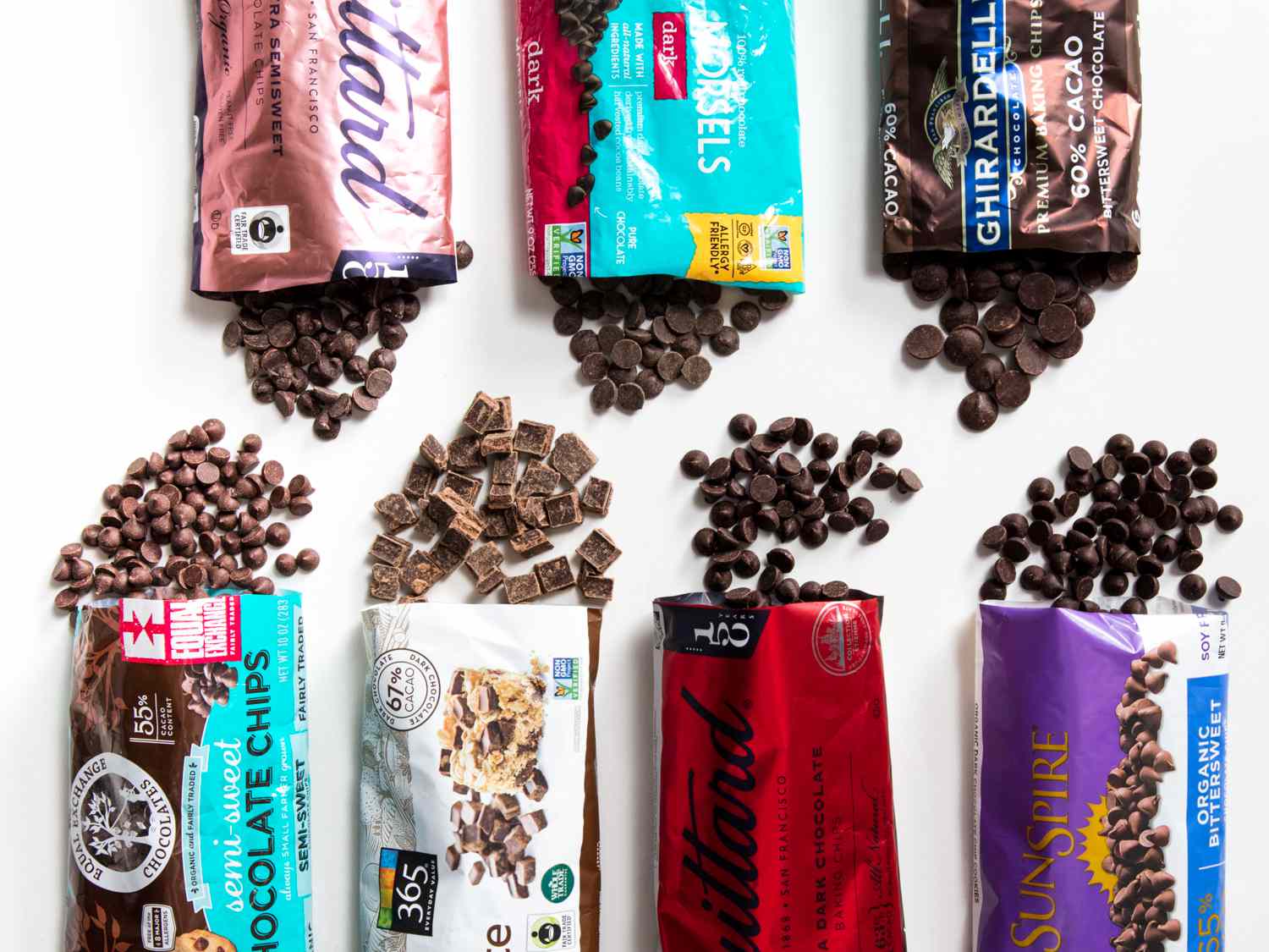 Six packets of chocolate chips opened on a white surface, spilling some of its content. The brands from top left to bottom right are: Guittard 55%, Enjoy Life, Ghirardelli, Equal Exchange, 365 Whole Foods, Guittard 63%, and Sunspire. 
