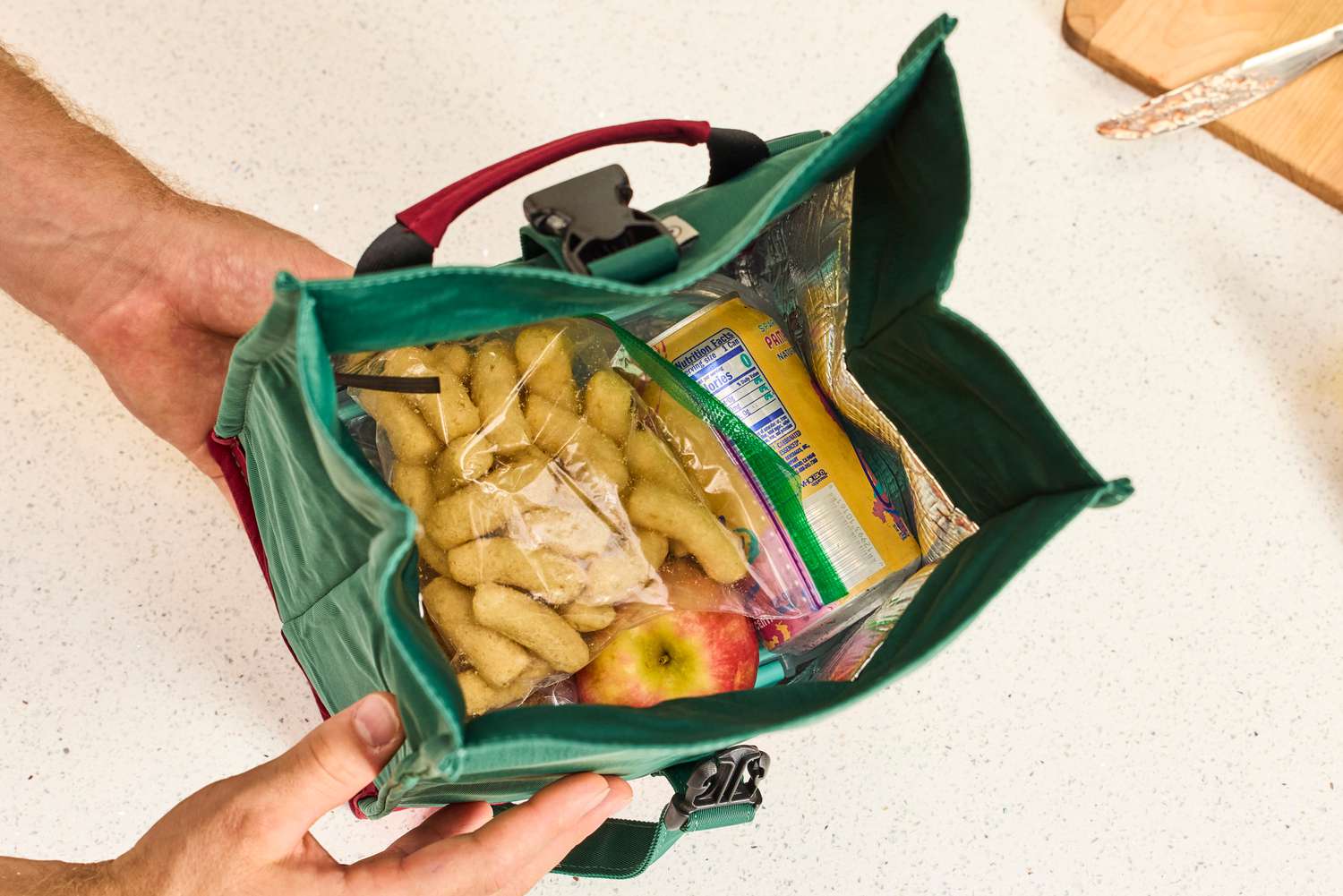 The inside of a green and red lunchbag, filled with an apple, a can of seltzer, and a bag of cheese puffs