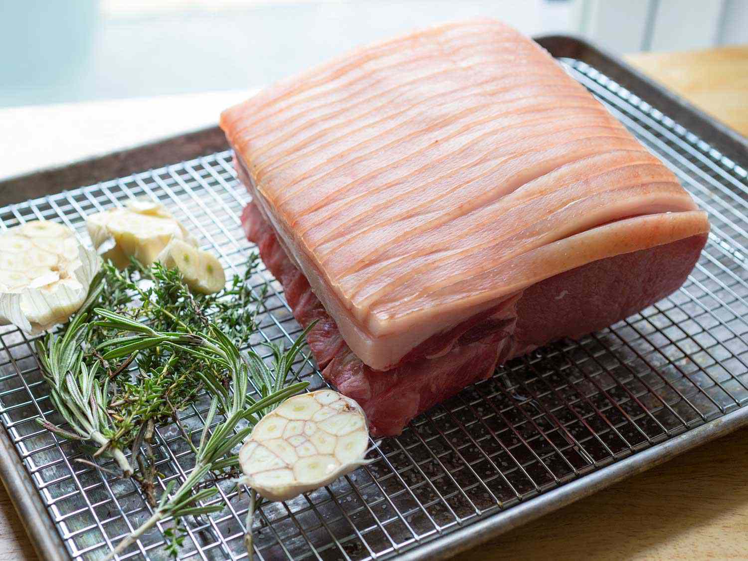 A whole, raw pork loin with a thick scored fat cap next to garlic and herbs