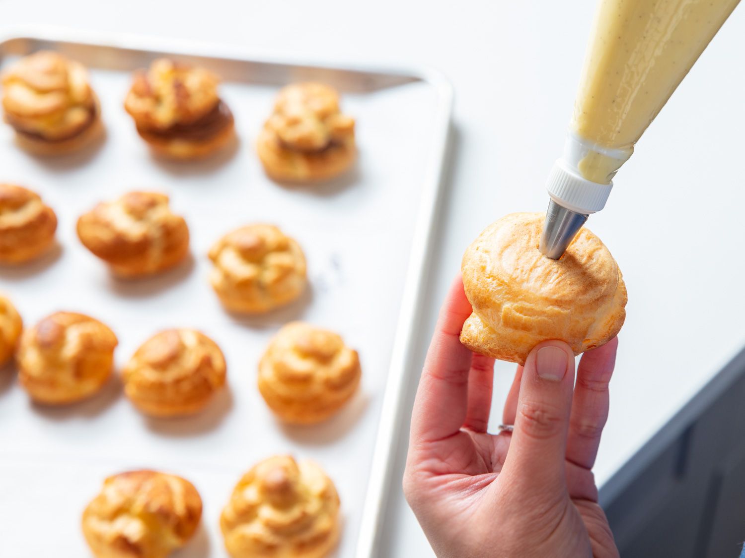 A cream puff being filled with cream with a baking sheet filled with puffs in the background.