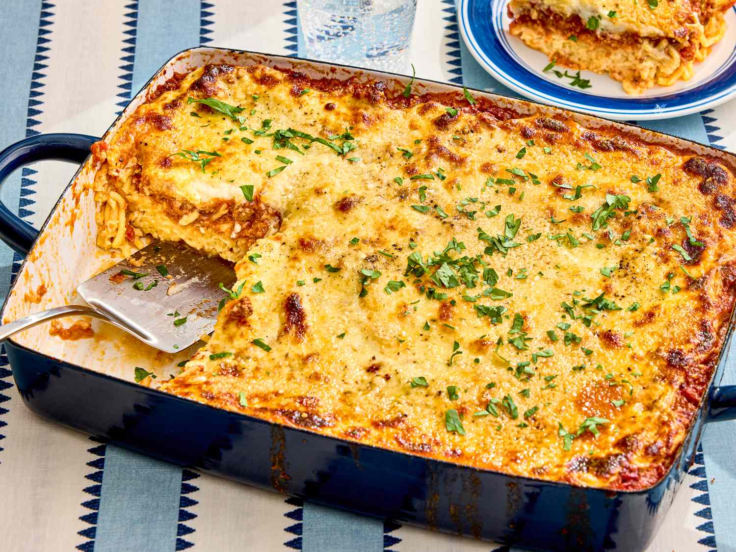A baked dish of spaghetti with a serving removed topped with herbs and cheese with a portion on a plate in the background