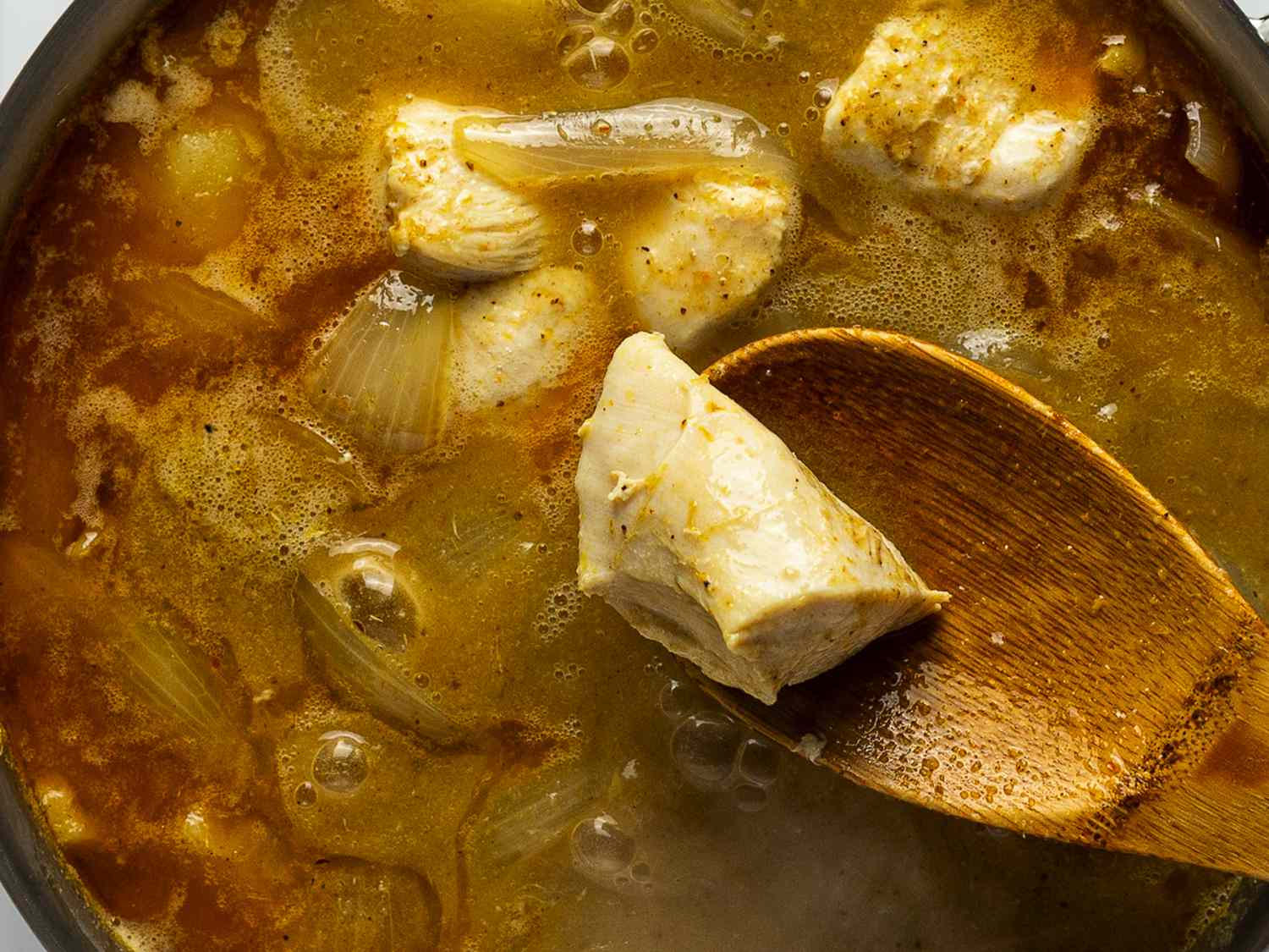 A close-up shot of the contents of the saucepan after the chicken has been added. A wooden spoon is lifting a piece of chicken out of the pan to demonstrate its cooked color and texture.