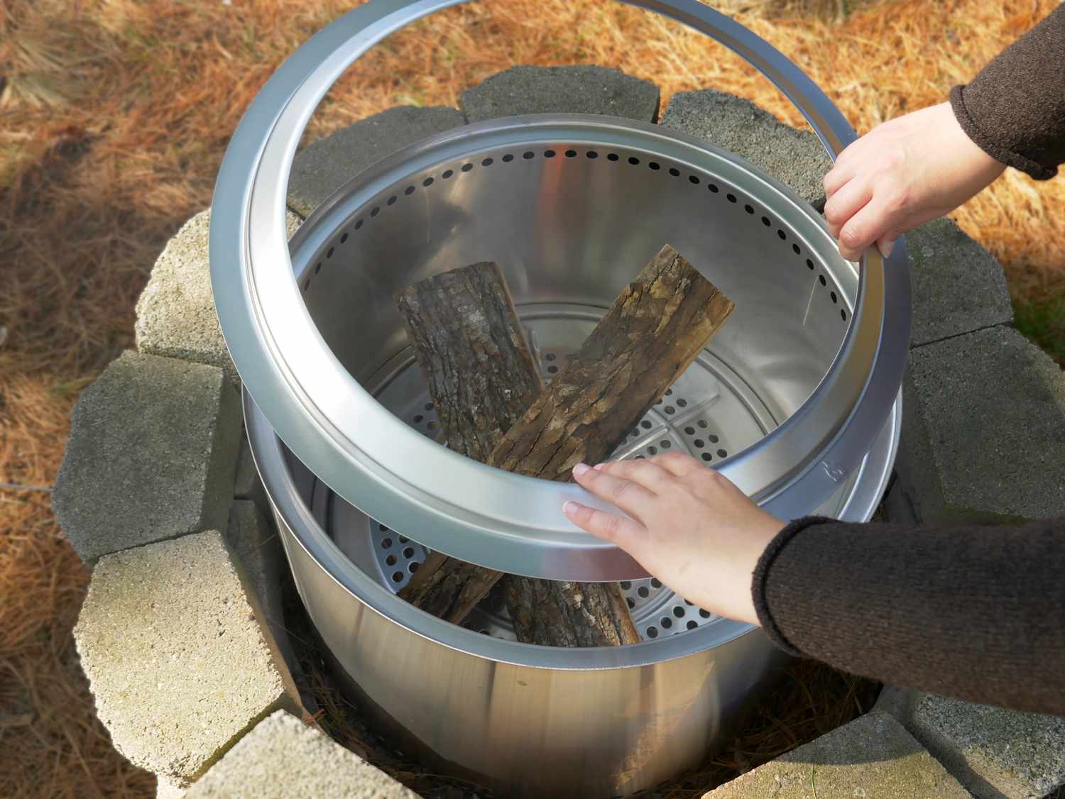 A person setting a ring of metal on top of a fire pit with wood inside.