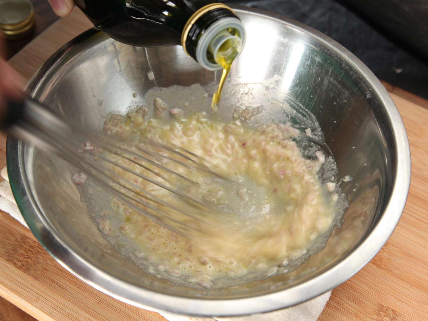 Process shot of making vinaigrette: olive oil is slowly drizzled into a mixing bowl as garlic and lemon juice are furiously whisked.