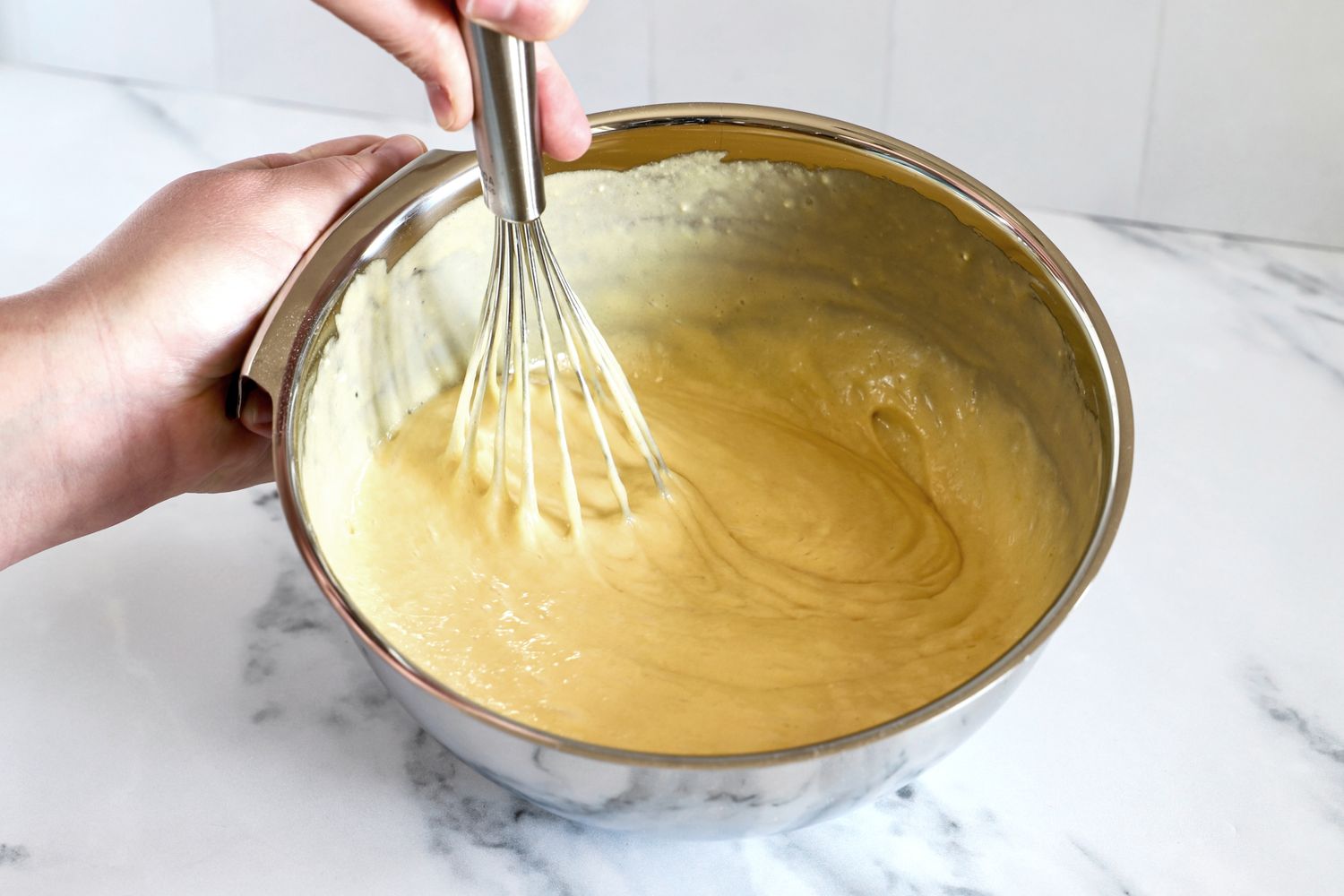 Hands whisking batter in an All-Clad mixing bowl on a marble countertop