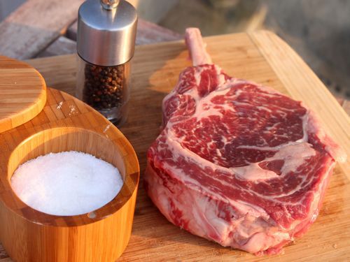 Thick marbled raw steak sitting on cutting board next to salt container and pepper shaker