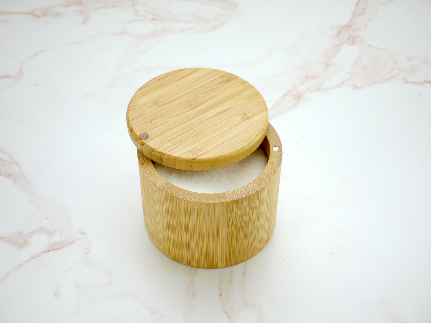 bamboo salt cellar with salt inside, lid slightly ajar, on a pink and white marble countertop