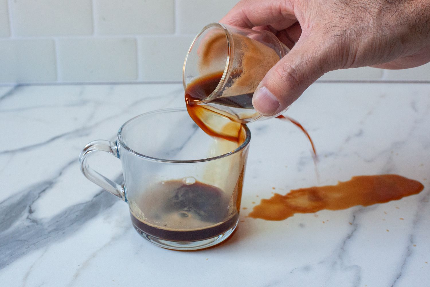 A person pouring espresso into a mug and it spilling on a marble countertop.
