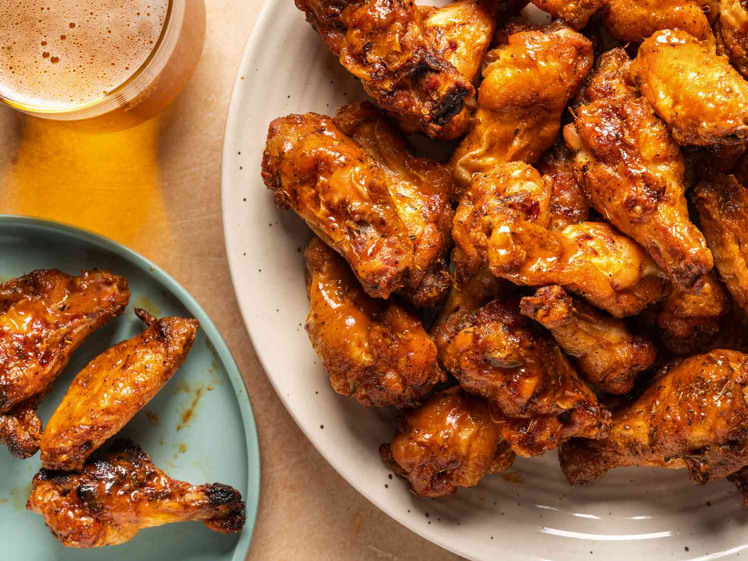 A large plate of grilled cajun chicken wings on the right hand side of the image. On the left side is a smaller blue plate with three chicken wings on it and a glass holding a glass of a golden-colored beverage.