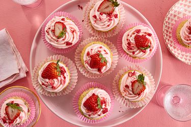 overhead shot of strawberry cupcakes on a pink tray and pink surface. with 2 on trays on the corners