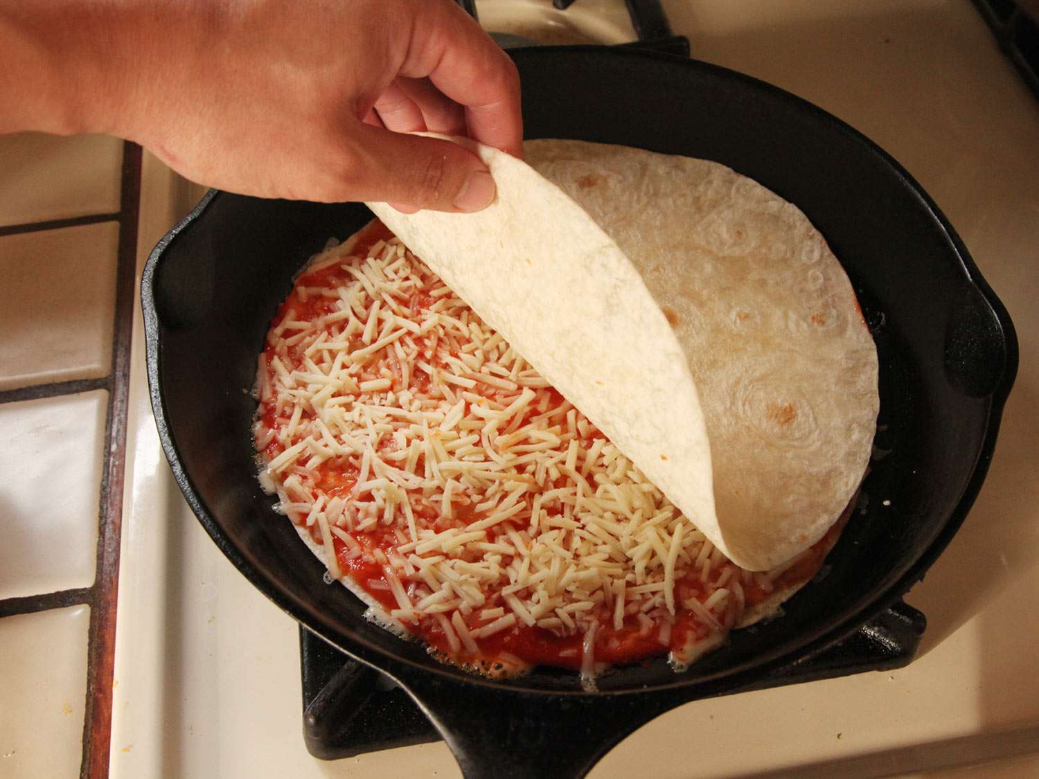 Topping off assembled cheese pizza layer in cast iron skillet with a second flour tortilla.