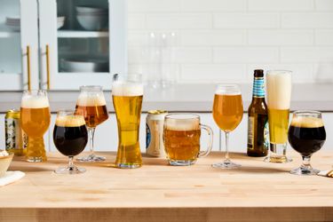 Numerous beer glasses on a kitchen countertop. Bottles and cans of beer are beside the glasses.