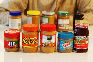 An assortment of different brands of peanut butters and jellies in jars on a table. 