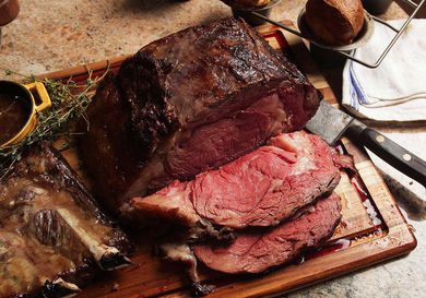 A whole cooked prime rib roast with two slices on a wooden cutting board with a knife, herbs, and sauce.