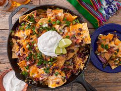 A skillet dish of loaded nachos garnished with lime wedges and sour cream; a small plate with a serving, on a wooden table