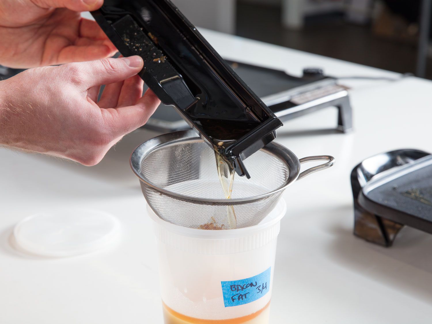 Fat being poured from a grease trap through a fine-mesh strainer into a labeled plastic container.