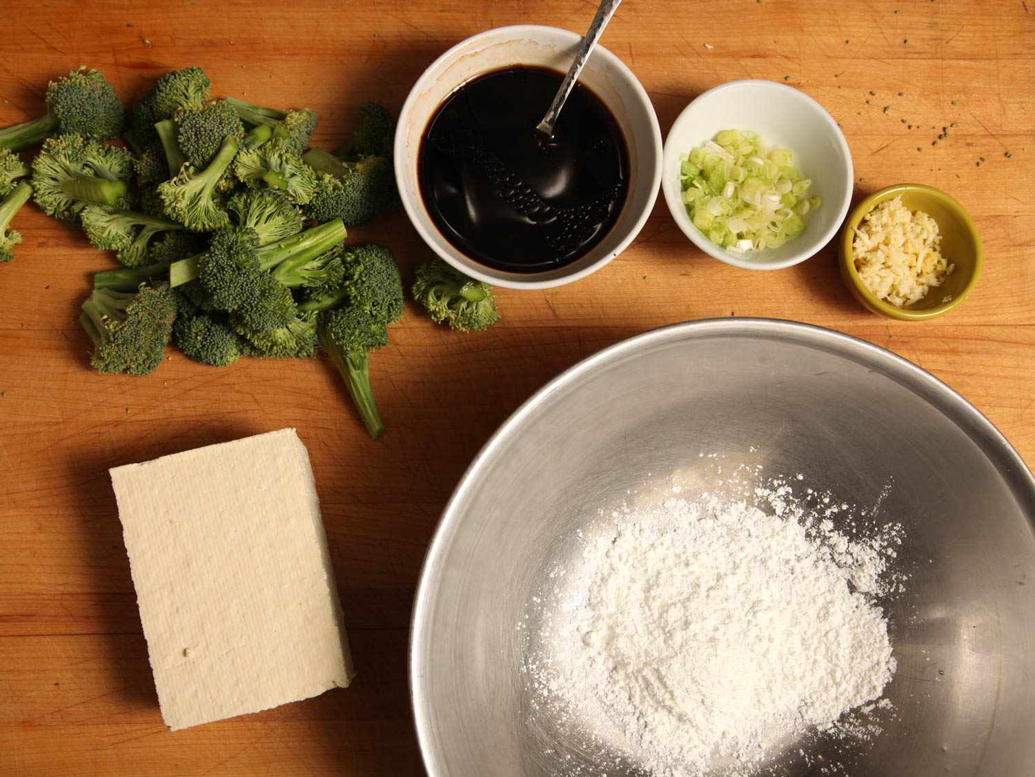 Ingredients on a wooden board: broccoli, soy sauce, sliced green onions, minced garlic, tofu, and cornstarch in a bowl. 