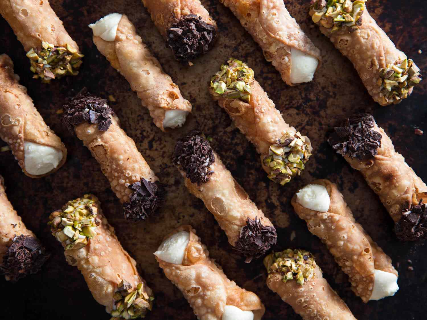 Overhead shot of cannolis lined up on a sheet pan. There are three kinds: plain, chocolate, and pistachio.