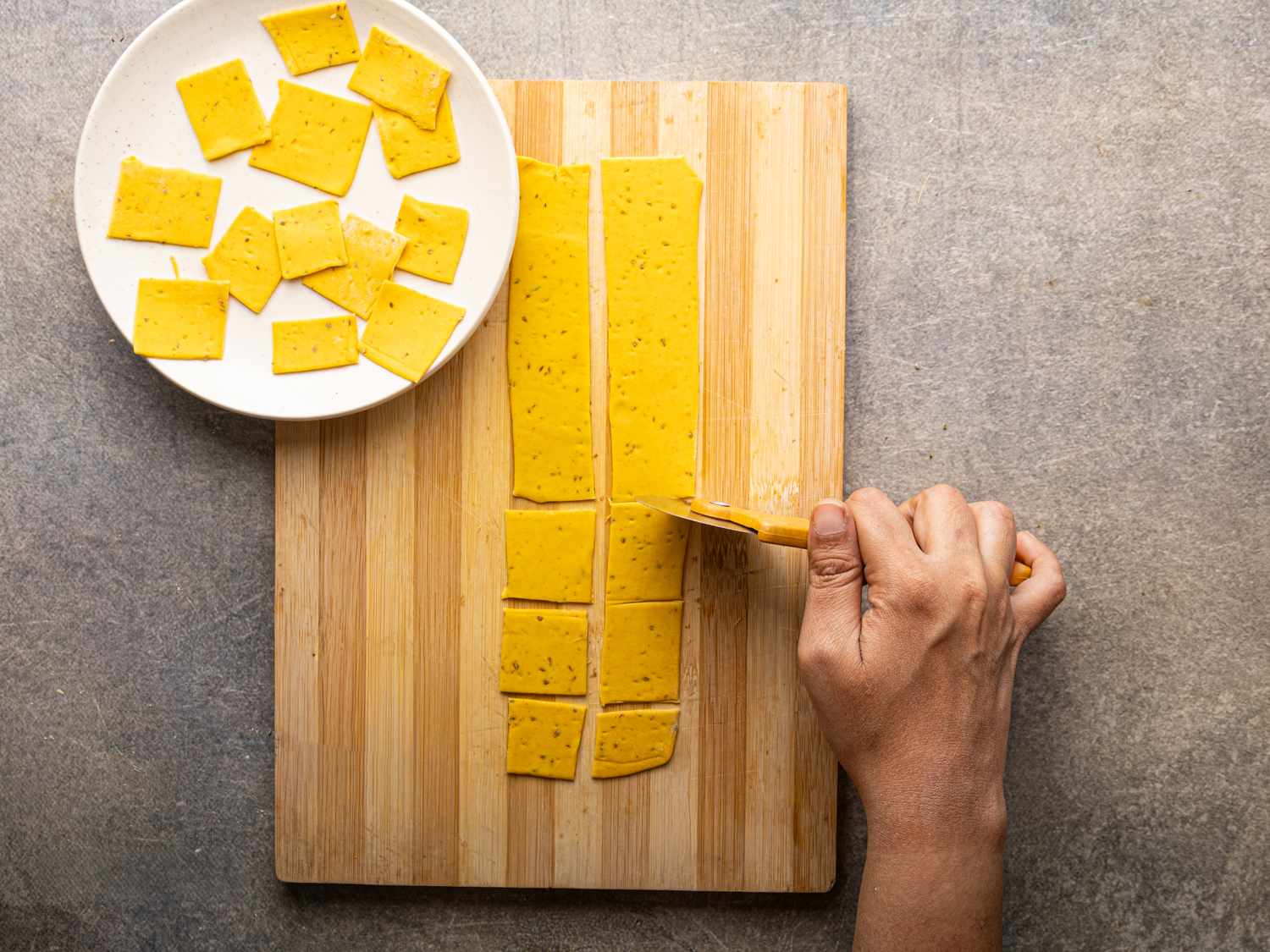 Person cutting yellow dough into squares on a wooden board with a plate of cut squares beside it
