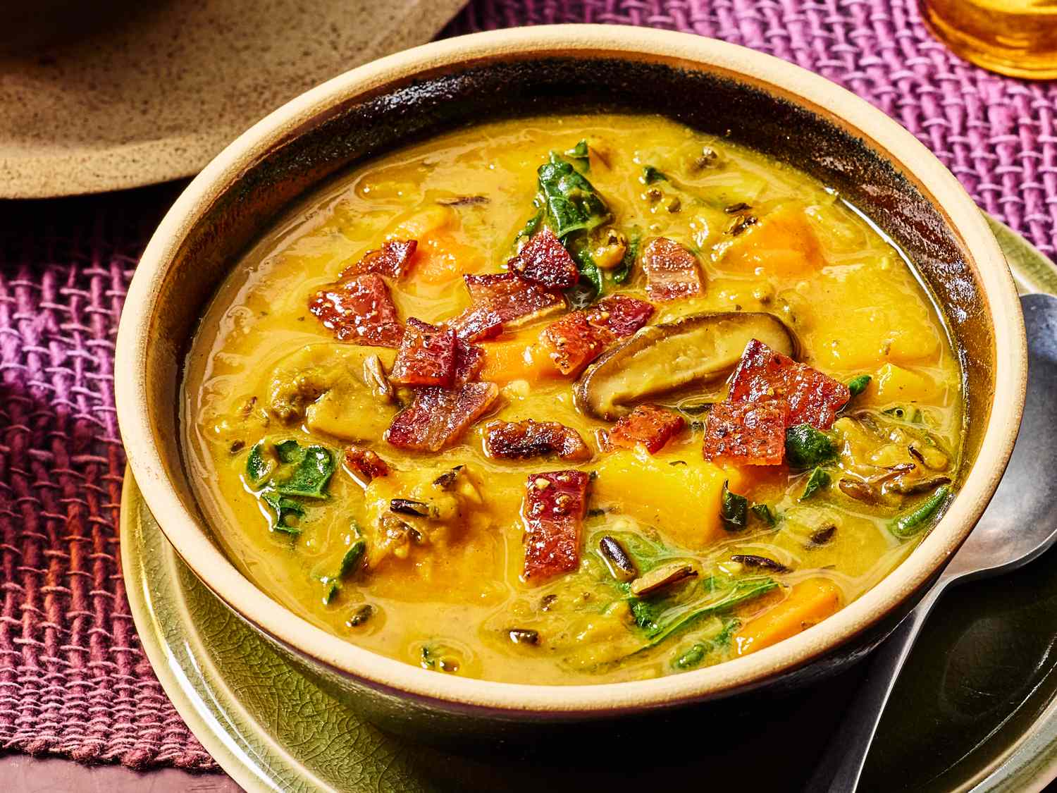 A bowl of wild rice and pumpkin soup garnished with vegetables and toppings placed on a plate with a spoon