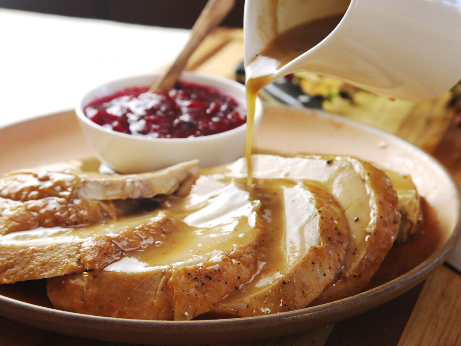 Gravy being poured from a small pitcher onto sliced turkey breast, on a plate with a dish of cranberry sauce.