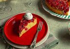 A slice of cranberry upsidedown cake on a red plate with a dollop of cream fork placed alongside the slice whole cake in background
