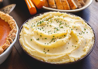 A bowl of mashed potatoes garnished with chives on a table with Thanksgiving dishes