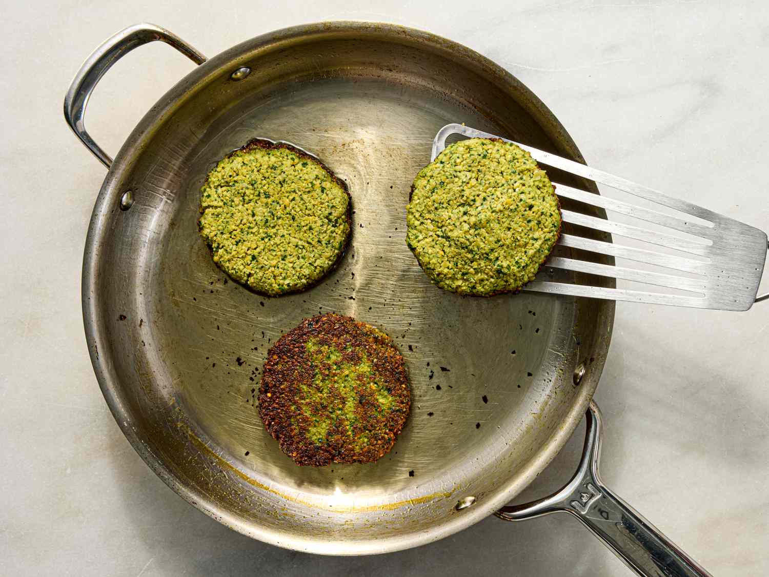 Falafel burgers being cooked in a pan with a spatula lifting one patty