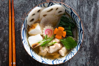A bowl of Japanese ozoni soup with sliced lotus root, chicken, mochi, and vegetables next to a pair of chopsticks.
