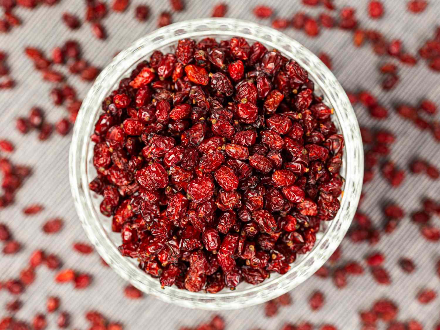 A bowl filled with dried red barberries surrounded by scattered barberries on a tabletop