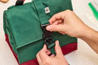 A person closing a buckle latch on a green and red roll-top lunch bag