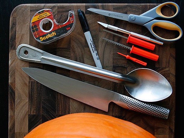 Various tools needed for carving a photograph into a jack-o'-lantern (Scotch tape, Sharpie marker, chef's knife, large spoon, carving picks, X-ACTO knife, scissors), arranged on a wooden cutting board