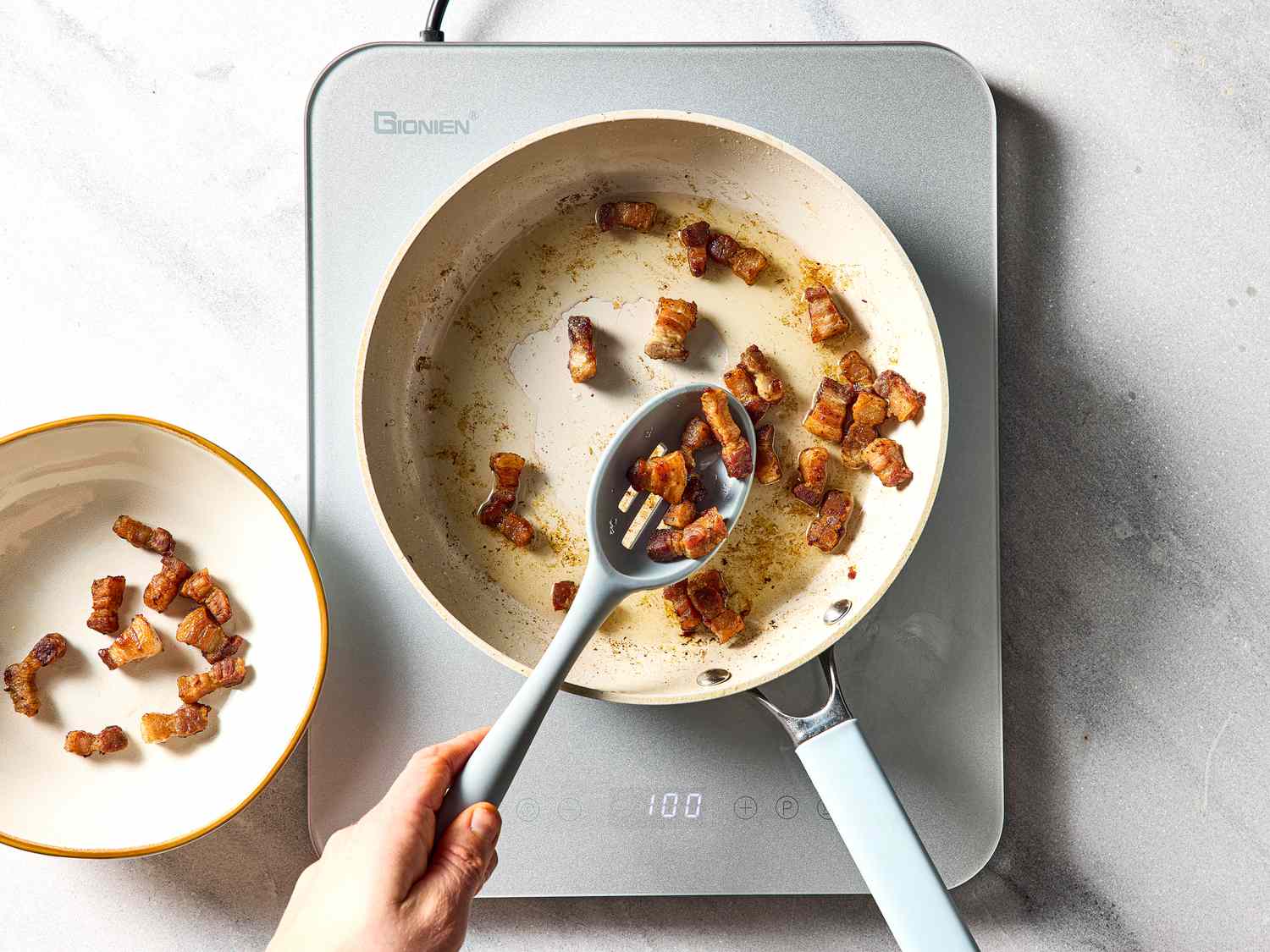 Cooking bacon pieces in a pan, a hand using a slotted spoon to transfer them to a plate