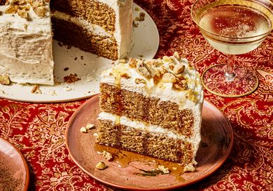 A slice of cake with layers of frosting and walnut garnish on a plate a partially sliced cake and a drink in the background