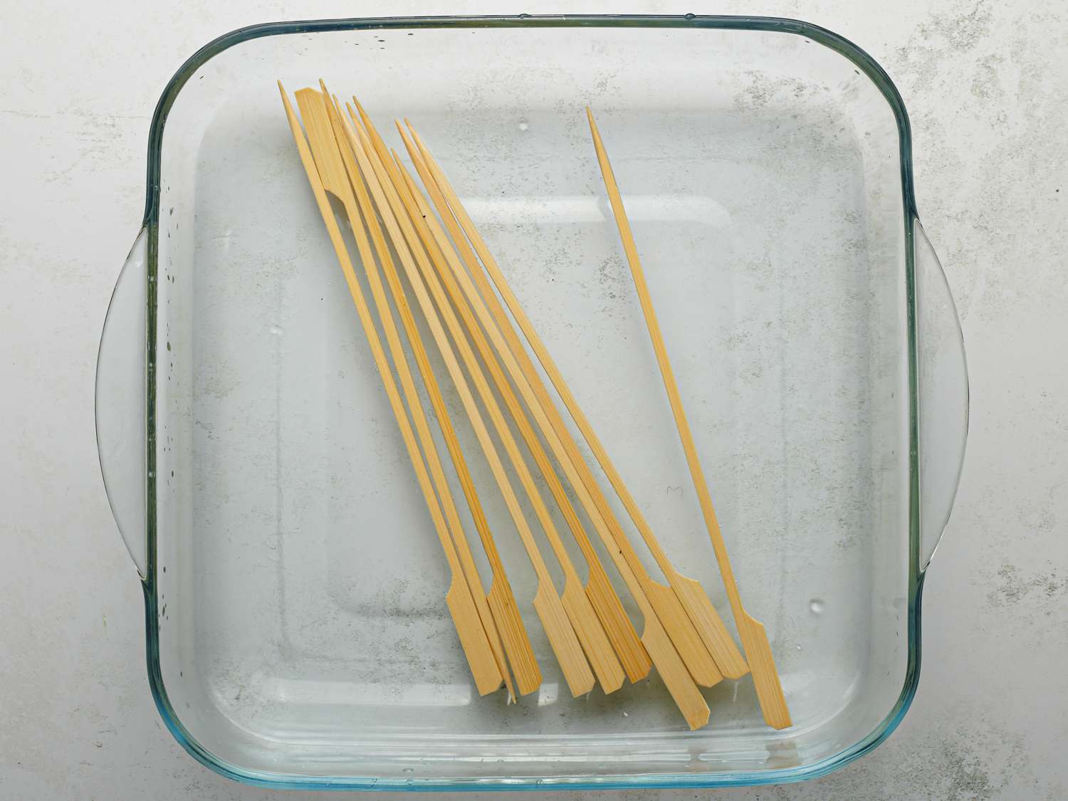 Bamboo skewers soaking in water inside of a square glass dish.