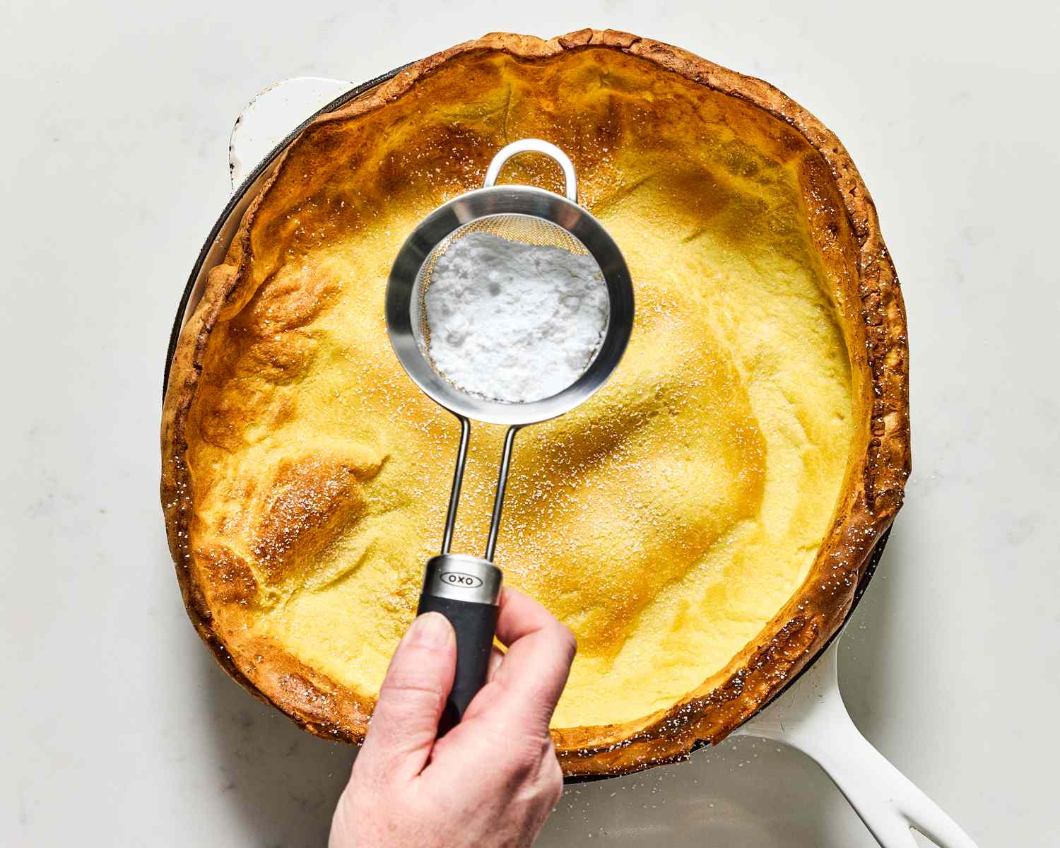 Person dusting powdered sugar onto a baked Dutch baby pancake