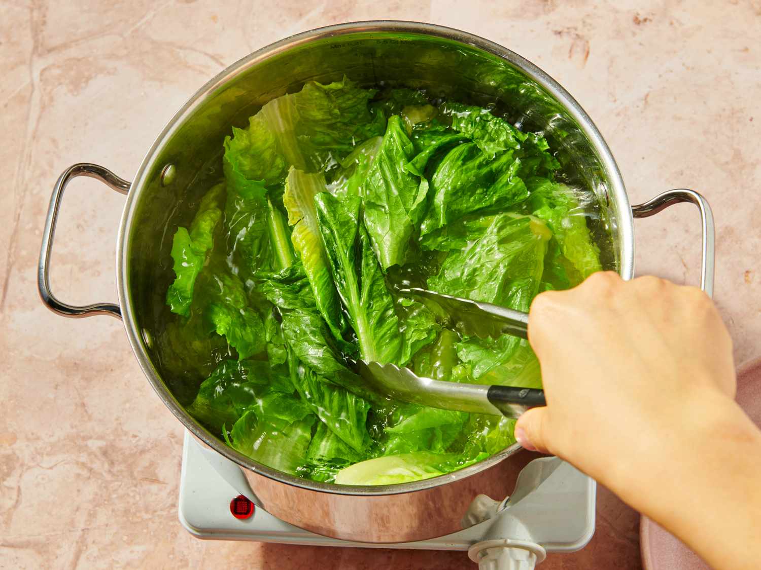 Lettuce being cooked in a pot of water with tongs held by a hand