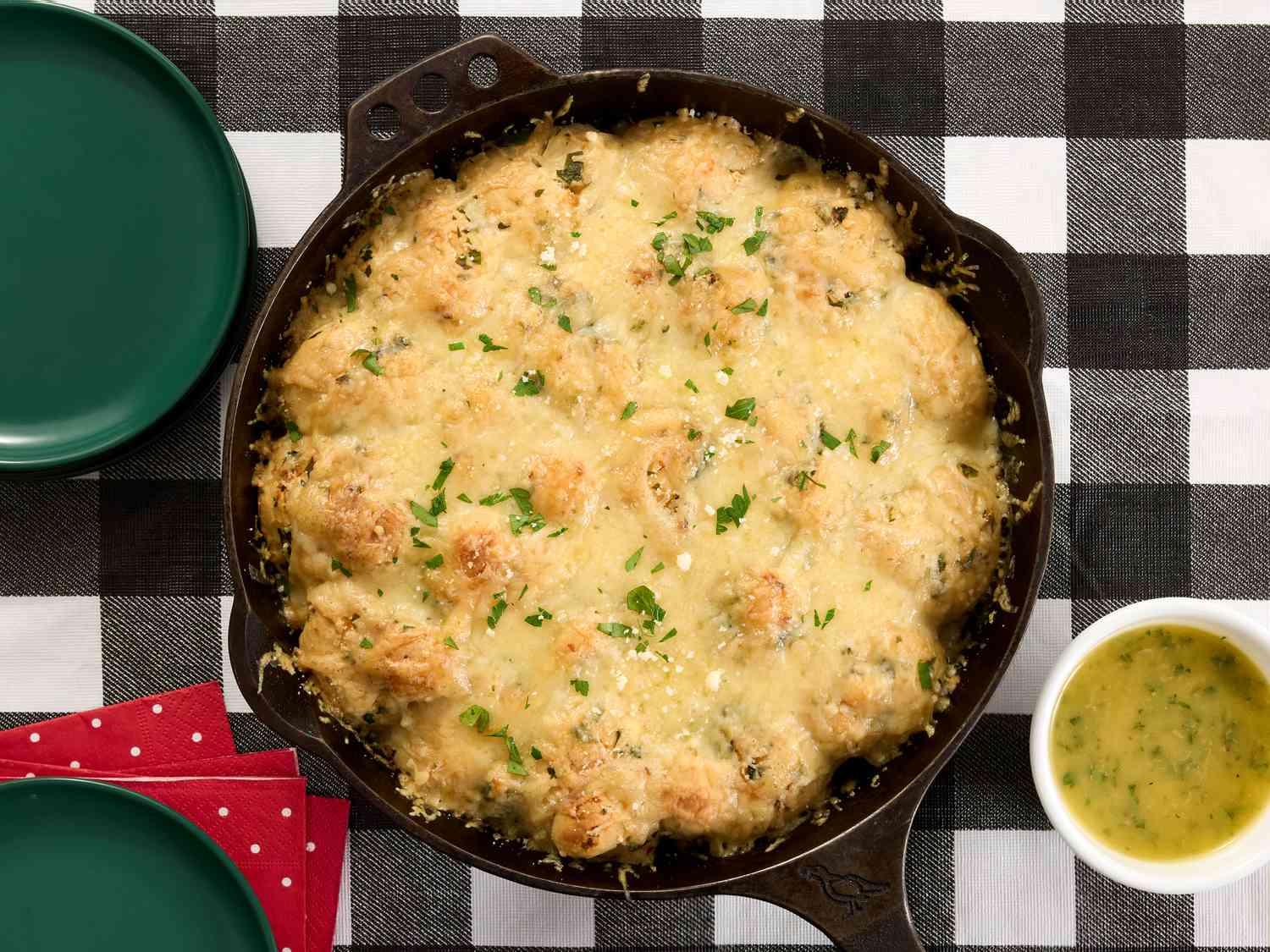 pullinh cheese garlic knot from cast iron pan and small dish of garlic butter on a black and white checkered fabric. 2 green plates and red napkin to the sides. 