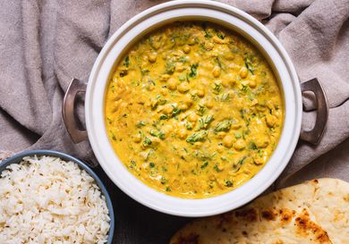 A double-handled serving crock with chickpea, coconut, and cashew curry. Some naan and a bowl of steamed rice are next to it. 