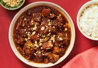 Overhead of stew in a bowl with small dish of peanuts in bowl to the side with red napkin and red background 