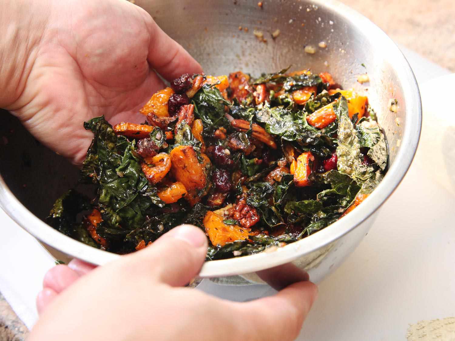 A hand tossing all of the ingredients for the salad in a mixing bowl, including roasted kale and squash, dried cranberries and pecans. 