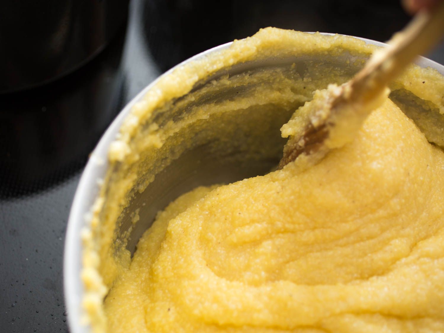 Scraping the sides of a pot filled with cooked polenta with a wooden spoon.