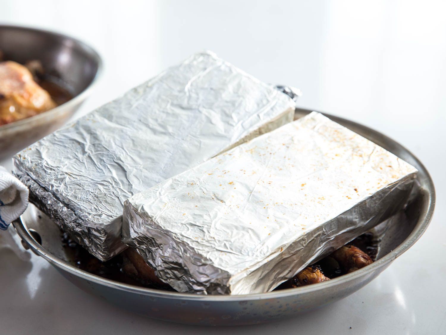 Two foil-wrapped bricks covering chicken in a metal skillet.