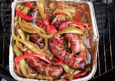 Overhead view of a finished batch of grilled Italian sausage with peppers and onions, piled in a disposable aluminum pan on a grill.