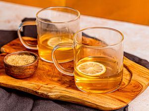 Two hot toddies served on a wooden board with lemon slices, next to a small bowl of demerara sugar